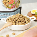 白色陶瓷碗裝滿方形凍乾幼貓主食餐，帶有淺色木勺，背景有生雞胸肉和切半煮蛋.