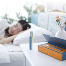 藍色玻璃睡眠噴霧瓶置於白色桌面，旁有橙色禮盒和一瓶粉色花瓶，背景模糊床上安睡女子.