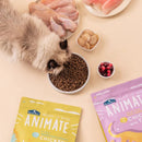 長毛貓正在吃碗中深色無穀貓飼料，旁邊有生雞肉、雞蛋和小碗莓果，兩包彩色Animate飼料包裝放於旁邊.